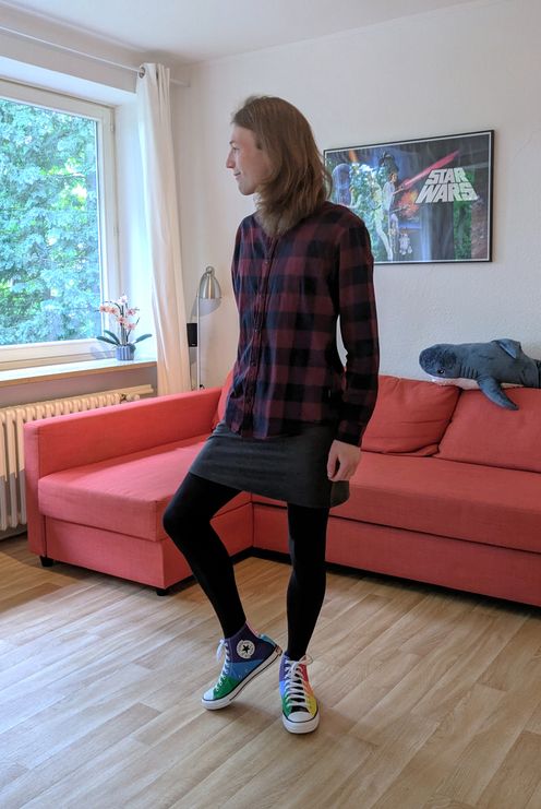 Photo of Fynn Ellie standing in a living room in front of a red couch with a Blåhaj plush shark. They wear a dark red and black checkered shirt, a dark gray skirt, black tights, and rainbow pride flag colored sneakers. They look to their right out of a window and their right knee is slightly angled upwards.