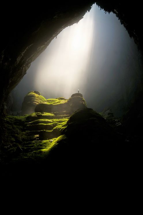 Lonely person standing on moss-covered rocks in a beam of light in a huge cavern.