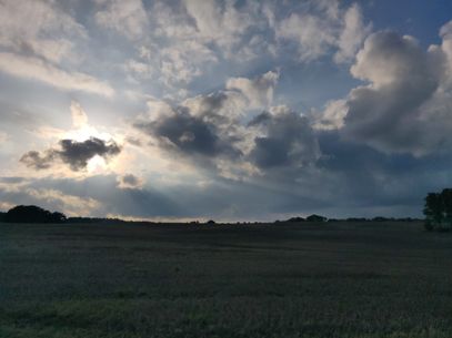 Photo at dusk: the landscape is already quite dark, a harvested field stretches to the horizon. A few dark trees are visible against the sky.
There is still some blue in the sky, but most of it is covered by heavy clouds.
Towards the left, a smaller cloud hangs in front of the sun, creating long godrays that streak to the right.
