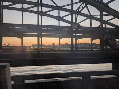 Through a steel truss bridge the orange hue of a sunset is visible, highlighting the skyline of the Port of Hamburg and HafenCity district. Underneath the bridge the calm water of the Elbe river is illuminated by the last rays of the setting sun.