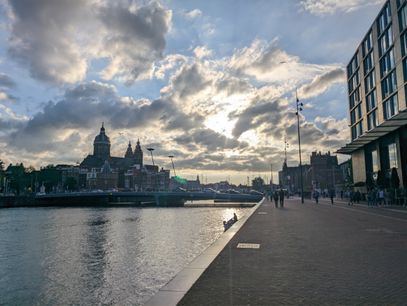 Evening sun peeking through some clouds over a canal in Amsterdam. Bridge over the water in the center. Medium and large sized buildings in the distance to the left and right. A lonely boat is floating on the canal.