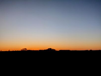 Photo of a sunset. A pitch black landscape. A few trees peak above the horizon in the far distance. The sky is a smooth cloud-less gradient from grayish blue to shiny orange at the horizon.