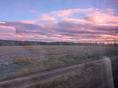 Photo of a sunset, taken from a moving train.
In the foreground is a harvested field that is quite blurry from the fast motion close to the camera but gets gradually clearer as it reaches the horizon.
There the field meets a line of trees that spans the entire width of the visible horizon.
The sky is a dark blue with some clouds lot red by the setting sun.
